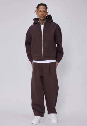 Homme avec des tatouages et des dreadlocks portant un sweat à capuche marron foncé zippé, un pantalon large assorti, une chemise blanche et des baskets blanches, debout sur un fond uni.