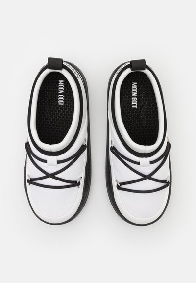 Pantofi slip-on alb și negru cu o talpă interioară texturată de culoare neagră, având curele elastice încrucișate, vârf rotund și o bandă de tragere la călcâi.