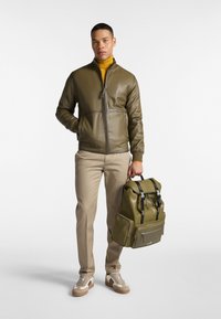 Olivgrüne Leder-Bomberjacke, beigefarbene Hose, hält einen passenden olivgrünen Rucksack mit schwarzen Trägern. Die Sneaker haben ein creme- und braunes Design.
