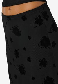 Svart skjørt laget av glatt stoff med et blomsterpattern med teksturerte, mørke fløyelsblomster. Midjebåndet er elastisk for en behagelig passform.