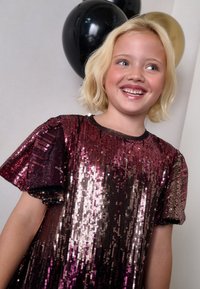 Spīdīgu kleita ar gradientu no tumši rozā līdz gaiši rozā krāsai, ar īsām piedurknēm un melnu apmali. Fonā melni un zelta baloni.