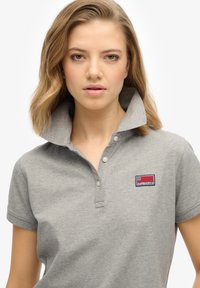 Grå polo-shirt lavet af bomuld, med en foldet krave, to knapper og et blåt og rødt broderet logo på venstre bryst.