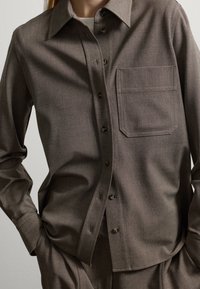 Persona che indossa una camicia button-up color taupe e pantaloni abbinati, con le mani nelle tasche, mostrando i dettagli della tasca anteriore e del colletto.