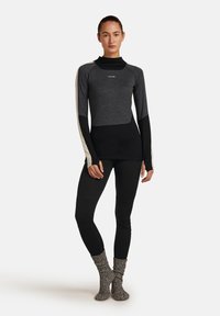Vrouw staat gekleed in een aansluitend zwart-grijs langwerpig shirt met duimopeningen, zwarte leggings en betegelde grijze sokken tegen een witte achtergrond.