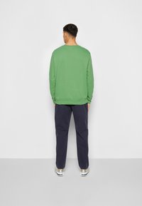 Homme de dos portant une chemise à manches longues verte, un pantalon bleu marine foncé et des baskets blanches sur fond blanc.