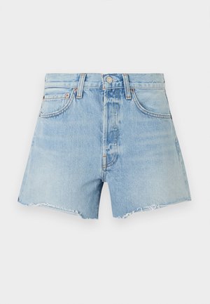 Femme portant un haut sans manches côtelé beige, un short en jean bleu clair effiloché et des sandales à talons noires, debout devant un fond blanc.