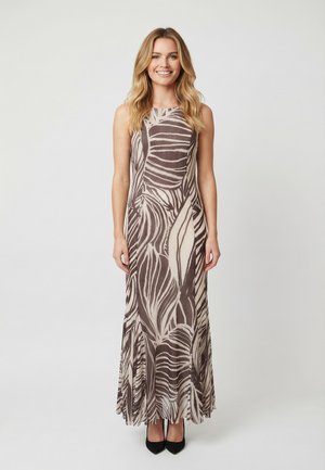 Mujer sonriente con cabello largo y rubio, vestida con un vestido sin mangas hasta el suelo con patrones abstractos en marrón y beige, y tacones negros.