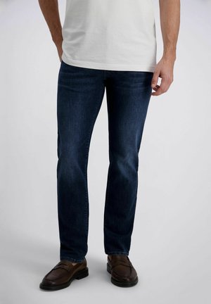 Mann trägt dunkelblaue Jeans mit geradem Bein, braune Slipper und ein weißes kurzärmeliges Hemd, steht vor einem einfarbigen Hintergrund.