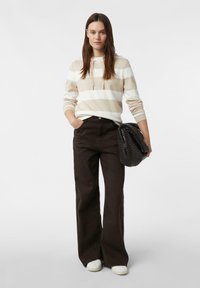 Beżowo-biała bluza w paski z kapturem i ściągaczem, zestawiona z czarnymi spodniami o szerokich nogawkach i wysokim stanie. W jednej ręce trzymana jest czarna torebka o fakturze.