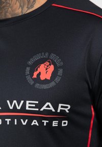Zwart sportshirt met rode accenten. Bevat een cirkelvormig logo met een gorillagrafiek en tekst. Materiaal lijkt glad en ademend.
