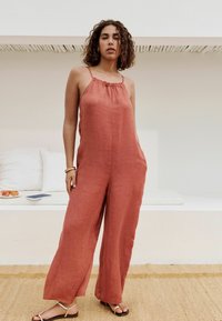 Jumpsuit od lanenog materijala u boji hrđe s nabranym izrezom i širokim nogavicama, uparen s minimalističkim sandalama, postavljen protiv neutralne pozadine.