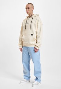 Felpa crema con logo nero, tasca kangaroo, cappuccio con coulisse; abbinata a jeans azzurri chiari e sneakers bianche. Stile semplice e taglio rilassato.