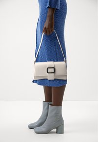 Borsa beige con una fibbia nera quadrata, tracolla e texture liscia, abbinata a un vestito blu a coste e stivali grigio chiaro.