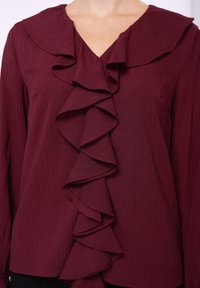 Burgundy blouse featuring a pronounced ruffle detail along the placket, textured fabric, and a wide collar with a v-neck design.