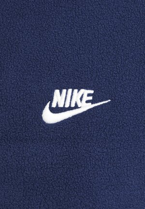 Granatowy materiał polarowy z wyszywanym białym logo "Nike" oraz detalem w kształcie łuku. Miękka tekstura z wzorzystym tłem.