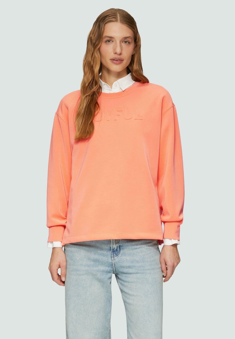 Koraalkleurige sweatshirt met een ronde hals, verlaagde schouders en reliëftekst "JOYFUL". Gedragen over een witte blouse en gecombineerd met lichtblauwe jeans.