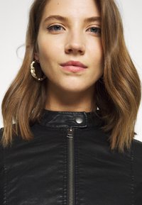 Black leather jacket with a high collar and silver zipper. Model wears textured gold hoop earrings. Close-up view of face and neck.