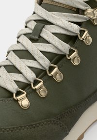 Botte de randonnée verte olive avec un dessus en tissu texturé, oeillets en métal doré et lacets gris clair avec un motif subtil.