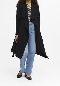 Langer schwarzer Cardigan mit offener Vorderseite, geschichtetem Design, getragen über einem gestreiften Oberteil und hellblauen Schlagjeans. Glattes Material, lässiger Stil.