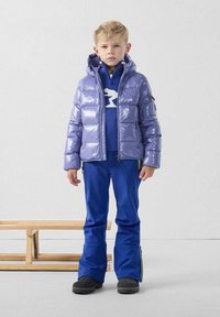 Chaqueta acolchada morada con acabado brillante, suéter de punto azul con un diseño blanco y pantalones azules. Botas negras completan el conjunto.