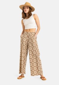 Pantaloni evazați cu model maro și design floral alb, purtați cu un top sans manches alb și o pălărie din paie, asortați cu sandale maronii.