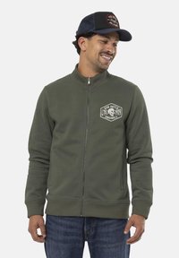 Sweatshirt zippé vert en matériau doux, présentant un patch logo rond sur la poitrine. Associé à un jean bleu, affichant une coupe décontractée.