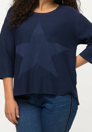 Femme portant un pull bleu marine avec un grand motif étoile sur le devant, associé à un jean bleu.