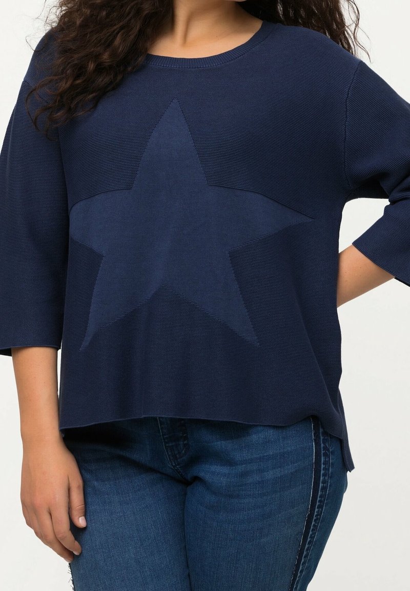 Femme portant un pull bleu marine avec un grand motif étoile sur le devant, associé à un jean bleu.