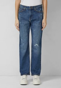 Niebieskie dżinsy z prostą nogawką, przetarciami oraz klasycznym fasonem z pięcioma kieszeniami. Noszone z białymi trampkami.