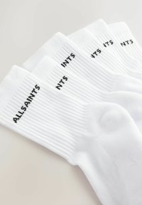 Chaussettes blanches côtelées avec un texte noir indiquant "ALLSAINTS" et "NTS". Design simple avec des poignets renforcés et une longueur classique à la cheville.