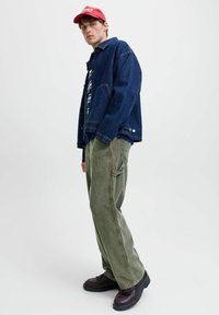 Blå denimjakke, stripete skjorte, olivengrønne kordfløyelsbukser og brune sko. Kappen er rød med hvit tekst.