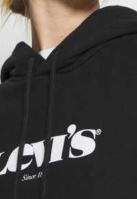 Ingrandimento di una persona che indossa un hoodie nero di Levi's con testo del logo bianco e lacci attorno al collo.