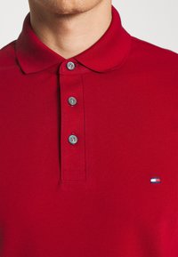 Polo rouge en tissu texturé, doté d'un col classique, de trois boutons argentés et d'un petit logo sur le côté gauche.