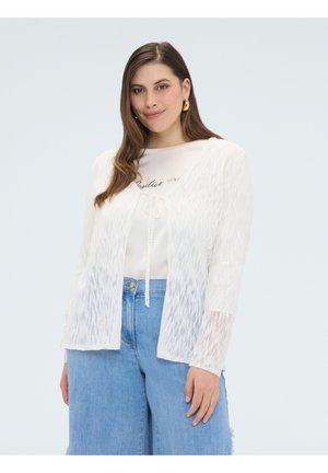 Vrouw met lang bruin haar die gouden oorringen draagt, een witte gestructureerde cardigan over een wit topje en blauwe high waist jeans tegen een effen achtergrond.