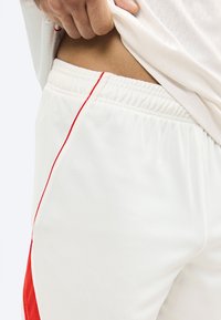 Χέρι τραβάει προς τα πάνω λευκό πουκάμισο, δείχνοντας το πλάι των λευκών αθλητικών σορτς με κόκκινη ρίγα και ελαστική μέση στον γοφό ενός ατόμου.