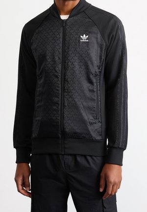 Mężczyzna noszący czarną kurtkę Adidas na zamek błyskawiczny z subtelnymi wzorami listka na przodzie i trzema paskami na rękawach.