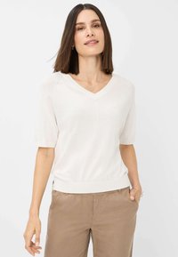 V-hals kortærmet sweater i lysebeige, blød tekstur, med ribbet kant og subtile syninger. Sammen med lysebrune bukser.