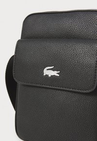 Sorte læder crossbody taske med en struktureret overflade, klaplukning og et sølvfarvet krokodillelogo-emblem på fronten.