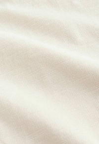 Nahaufnahme eines weichen, cremefarbenen Stoffs mit feiner Strickstruktur und sanften Falten, die subtile Schatten und Lichtreflexe erzeugen.