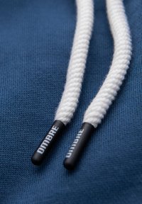 Vita vävda dragsnören med svarta plaständar märkta "UMBRO" på blått stickat tyg.