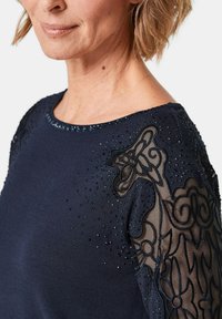 Marineblauer Pullover mit perlenbesetzten Verzierungen an den Schultern und zarten Spitzenakzenten an den Ärmeln. Weicher Stoff mit strukturierten Details.