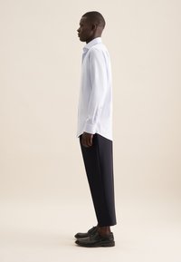 Camicia a maniche lunghe a quadri blu chiaro, abbinata a pantaloni neri su misura e scarpe nere, vista di lato su uno sfondo neutro.