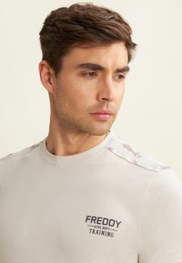T-shirt beige chiaro con colletto a girocollo, maniche corte e dettagli a camuflaggio sulle spalle. Logo stampato nero sul petto.