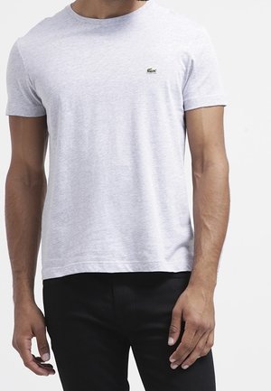 Mann trägt ein schlichtes hellgraues T-Shirt mit kurzen Ärmeln, auf der linken Brust ein kleines grünes Krokodil-Logo und schwarze Hose.