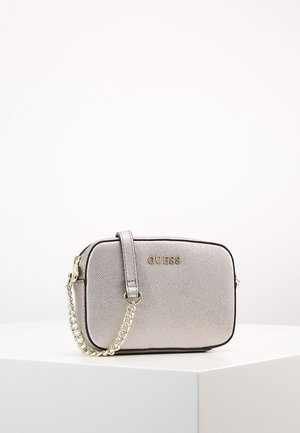 Petit sac à main rectangulaire argenté Guess avec une bandoulière chaîne dorée reposant sur une surface blanche sur un fond uni.