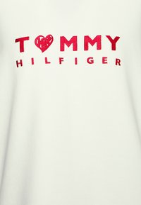 Biała koszulka z czerwonym haftowanym napisem "TOMMY" z symbolem serca, a następnie "HILFIGER," o gładkiej fakturze.