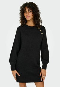 Zwart gebreide trui jurk met een ronde hals, lange pofmouwen en drie decoratieve gouden knopen op de linker schouder. Geweven stof.
