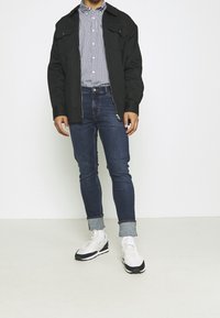 Czarna kurtka na zamek, niebieska koszula w kratę, ciemnoniebieskie jeansy slim fit, podwinięte mankiety oraz białe/czarne trampki z akcentami z siateczki i zamszu.