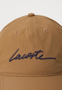 Chapeau marron en tissu léger, orné d'un logo "Laureate" brodé en bleu marine à l'avant. Texture lisse, forme structurée.
