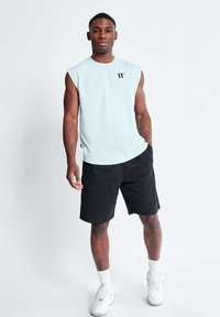 Camisa sin mangas de color azul claro con un logo negro, combinada con pantalones cortos negros y zapatillas blancas, de pie sobre un fondo claro.